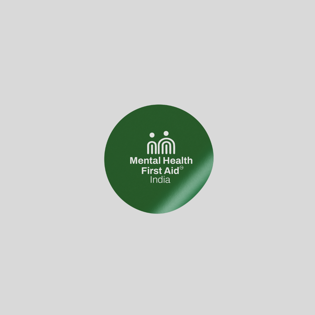 A circular dark green button badge with the white text 'Mental Health First Aid India' and a small white icon, centered on a light gray background.