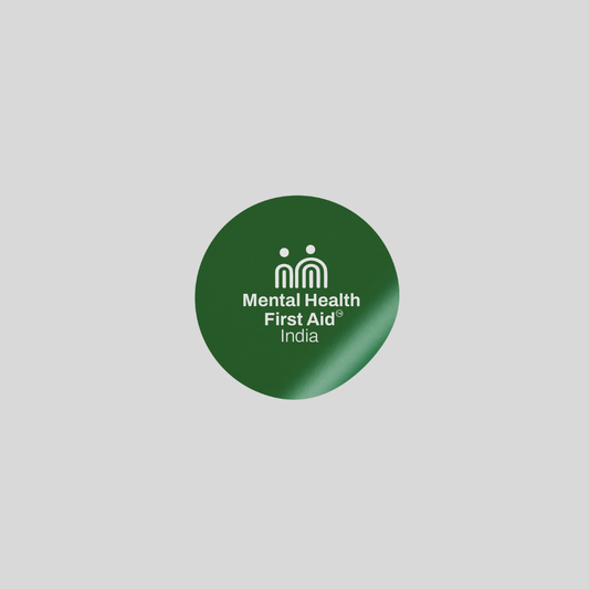 A circular dark green button badge with the white text 'Mental Health First Aid India' and a small white icon, centered on a light gray background.