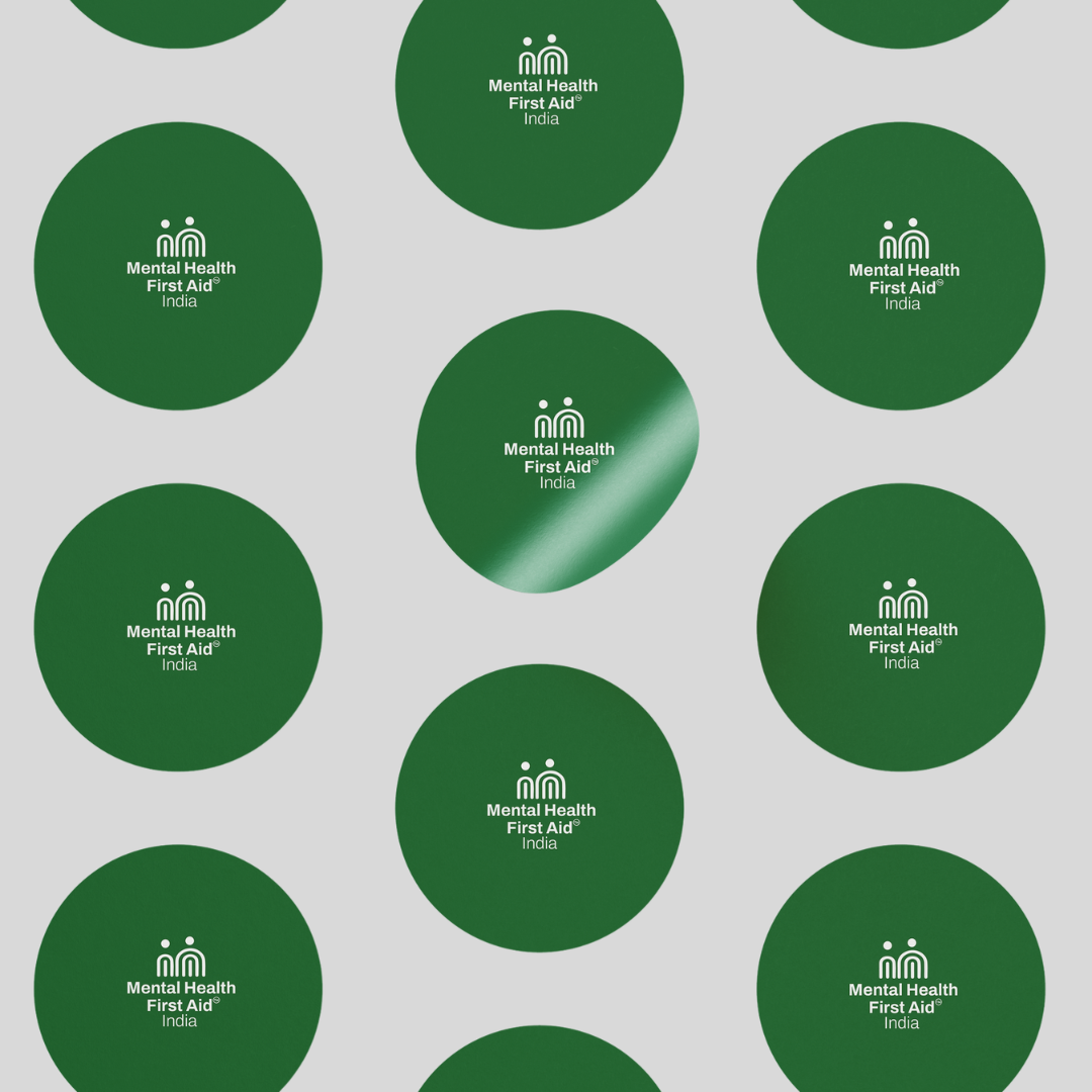 A repeating pattern of dark green circular stickers on a light gray background, each printed with the white text and logo reading 'Mental Health First Aid India'.
