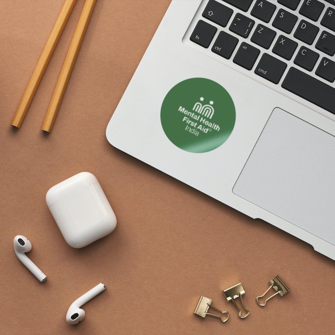 A green circular sticker with the text 'Mental Health First Step Pizza' and a white logo is shown on the corner of a silver laptop. The laptop is placed on a brown surface alongside wooden chopsticks, white wireless earbuds in their charging case, and gold binder clips.
