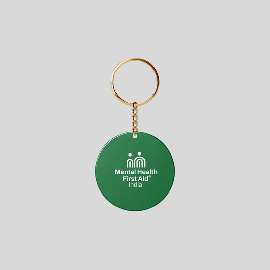 A green circular keychain with a gold metal key ring and chain, featuring the white printed text and logo for 'Mental Health First Aid India' on a light gray background.